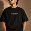 THE HOUSE TEE — INK — editorial front (model) Editorial lookbook mockup: model wearing the black THE HOUSE TEE — INK, front view with the small ivory LUXORIQ wordmark and IN EVERY THREAD on the chest
